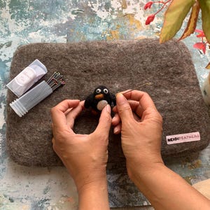 Puede incluir: Una persona fieltro un pingüino blanco y negro con una aguja y un trozo de fieltro marrón. El fieltro está sobre una superficie gris. La persona está usando una herramienta de fieltro de aguja para agregar detalles al pingüino.