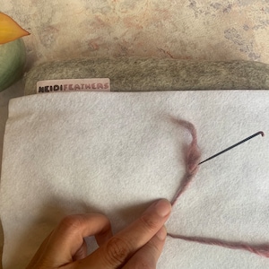 Puede incluir: Primer plano de una aguja de fieltro y un hilo de lana rosa sobre una alfombra de fieltro blanca. La aguja se utiliza para felpar la lana junta. La alfombra está sobre un trozo de fieltro gris con una etiqueta que dice "HeidiFeathers".
