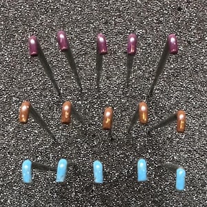 May include: Fifteen straight pins with rounded heads arranged in three horizontal rows. The top row features five dark purple heads, the middle row has five copper-colored heads, and the bottom row contains five light blue heads.
