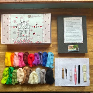May include: A craft kit with a pink box featuring a birdcage design and the text "heide feathers." The kit includes colorful wool roving, instructions, and tools for 2D picture making. The wool colors range from yellow to black.