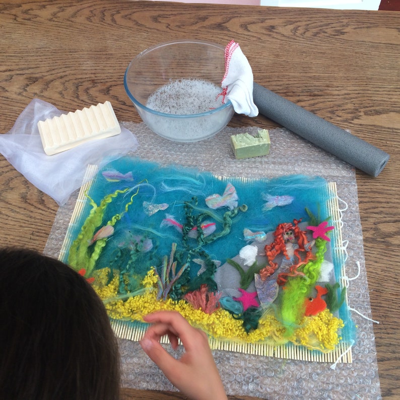 May include: A close-up of a needle felted mermaid scene with a mermaid, seaweed, and fish. The scene is on a bamboo mat and is being worked on by a person.