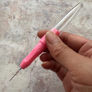 Può includere: Uno strumento ad ago rosa e trasparente con un ago lungo e sottile che si estende dall'impugnatura rosa. Lo strumento ha un cappuccio di plastica trasparente. Lo strumento è tenuto in una mano su uno sfondo screziato.