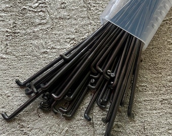 Puede incluir: Un paquete de ganchos de metal negro en un contenedor de plástico transparente. Los ganchos son todos del mismo tamaño y forma, y están dispuestos en una pila ordenada.