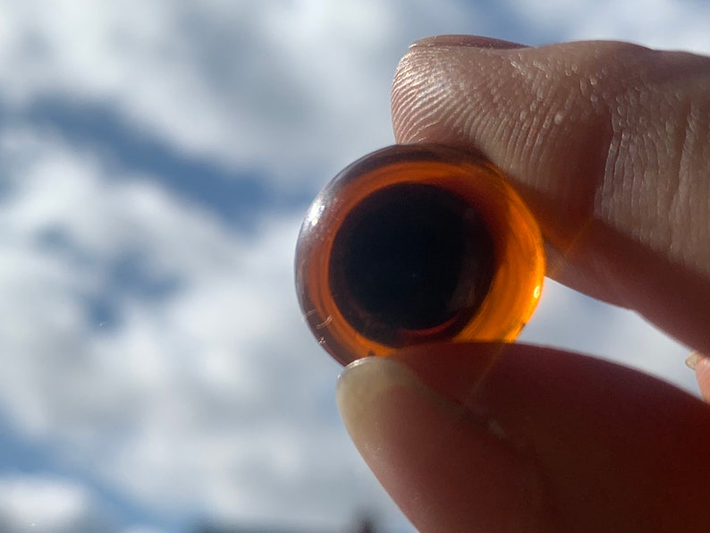 May include: A close-up of a round, amber-coloured glass eye with a black pupil. The eye is being held by a finger.