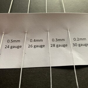 Może przedstawiać: Biała karta z pięcioma białymi drutami przymocowanymi do niej. Karta zawiera tekst: "0,6 mm gauge", "0,5 mm 24 gauge", "0,4 mm 26 gauge", "0,3 mm 28 gauge" i "0,2 mm 30 gauge".