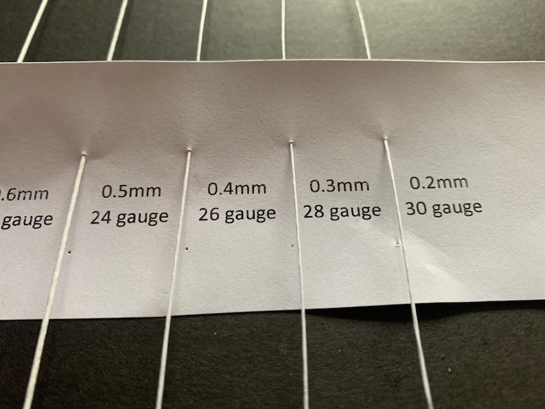 Może przedstawiać: Biała karta z pięcioma białymi drutami przymocowanymi do niej. Karta zawiera tekst: "0,6 mm gauge", "0,5 mm 24 gauge", "0,4 mm 26 gauge", "0,3 mm 28 gauge" i "0,2 mm 30 gauge".