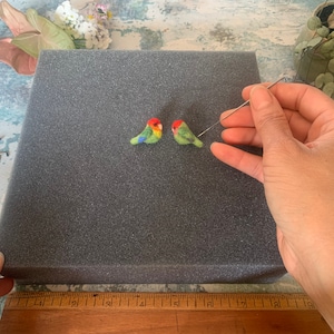May include: Two small felt birds, one green and red, the other green and orange, are being needle felted on a gray foam surface. A felting needle is held in the right hand of the person working on the birds.