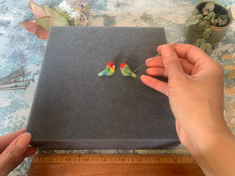May include: Two small felt birds, one green and red, the other green and orange, are being needle felted on a gray foam surface. A felting needle is held in the right hand of the person working on the birds.