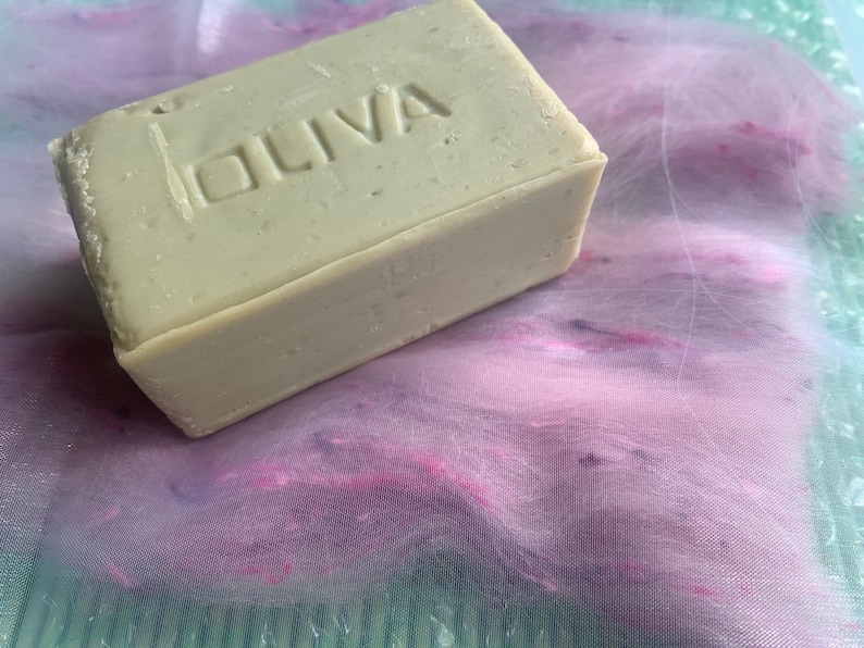 May include: A bar of olive oil soap, labelled "OLIVA", on a pink and purple fabric background.
