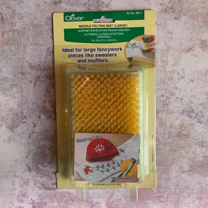 以下が含まれることがあります： プラスチックのパッケージに入った黄色のニードルフェルトマット。パッケージは緑色の背景に「Clover Needle Felting Mat (Large)」と「大きなファンシーワーク、セーターやマフラーなどに最適です」というテキストが書かれています。パッケージには、スカーフとキャップのデザインアイデアも示されています。