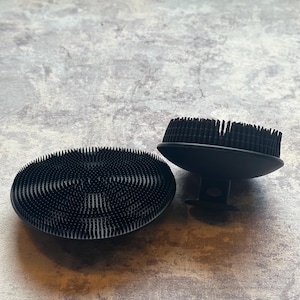 May include: Two black circular silicone body scrubbers featuring numerous short, flexible bristles on the top surface. One scrubber lies flat, while the other is positioned upright, revealing a small handle or grip on its underside for holding.