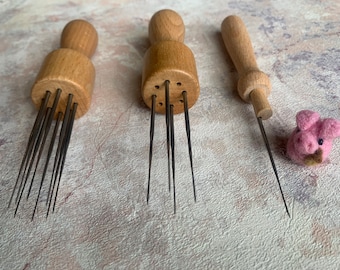 May include: Three wooden needle felting tools with metal needles. The tools have different needle configurations. A pink felt pig is in the background.