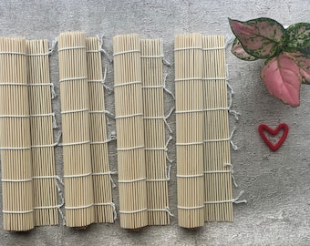 May include: Six natural bamboo sushi rolling mats with white string ties. The mats are laid out in a row on a gray textured surface.
