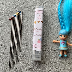 May include: A set of ten colorful, metal knitting needles in a clear plastic tube. The tube has a white label with the text "heidi" and a small bird illustration.