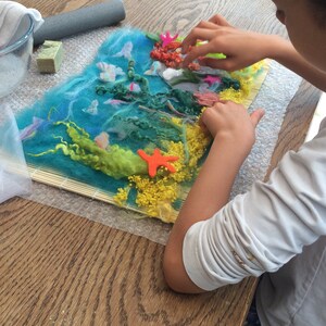 May include: A child is working on a felt art project. The project depicts an underwater scene with blue, green, and yellow felt. The child is adding orange and red felt pieces to the project.