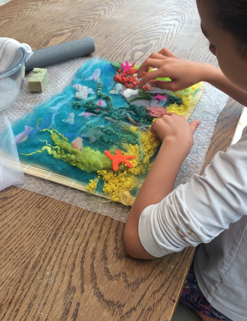 May include: A child is working on a felt art project. The project depicts an underwater scene with blue, green, and yellow felt. The child is adding orange and red felt pieces to the project.