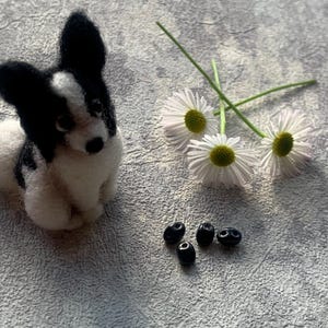 May include: A black and white felt dog figurine with a long tail, sitting on a gray textured surface. There are three white daisies and five black beads on the surface.