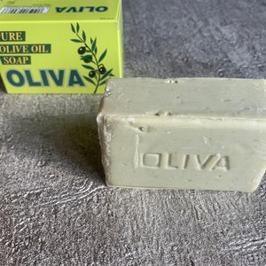 May include: A bar of light grey soap with the word "OLIVA" embossed on the front. The soap is sitting on a grey textured surface. A yellow box with the words "PURE OLIVE OIL SOAP OLIVA" is visible in the background.