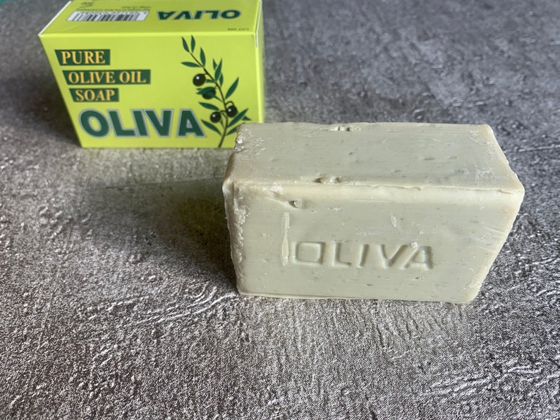 May include: A bar of light grey soap with the word "OLIVA" embossed on the front. The soap is sitting on a grey textured surface. A yellow box with the words "PURE OLIVE OIL SOAP OLIVA" is visible in the background.