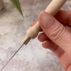 May include: A wooden needle threader with a long, thin metal needle. The threader has a wooden handle and a small metal loop at the end of the needle.