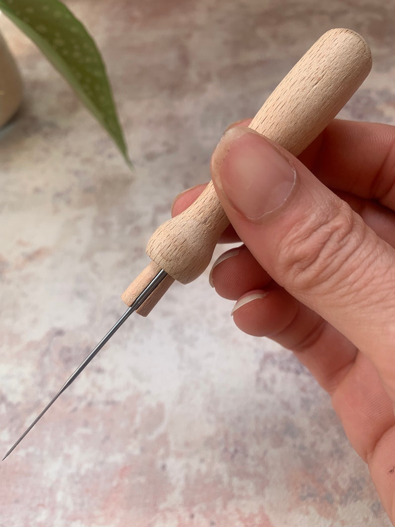 May include: A wooden needle threader with a long, thin metal needle. The threader has a wooden handle and a small metal loop at the end of the needle.