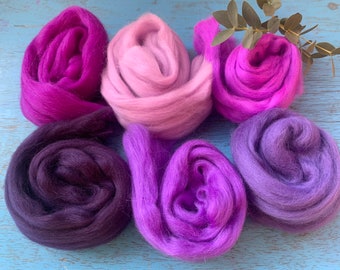 Puede incluir: Seis ovillos de lana en tonos de morado, rosa y lavanda. La lana está dispuesta en círculo sobre una superficie de madera azul.