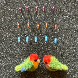 May include: Two needle-felted birds with red heads, green bodies, and blue wing accents positioned below fifteen sewing pins with metallic purple, copper, and light blue spherical heads arranged in three horizontal rows of five pins each.