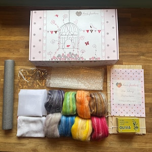 May include: A wet felting kit featuring a white box with a birdcage illustration and the text heidi feathers, eight bundles of colorful wool roving, a bamboo mat, a bar of Oliva pure olive oil soap, a white mesh sheet, and a gray foam roller.