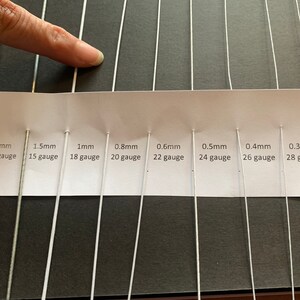 Może przedstawiać: Biała karta z rzędem dziesięciu białych metalowych drut&oacute;w o r&oacute;żnych grubościach. Druty są oznaczone średnicą w milimetrach i grubością. Karta leży na czarnej powierzchni.