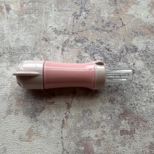 以下が含まれることがあります： ピンクとベージュのキッチンツール。円筒形のピンク色の中心部、ベージュ色の端、金属製のピンが付いた透明な長方形のセクションがあります。ツールには、片方の端に小さな翼のようなディテールがあります。