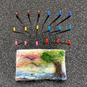 Könnte beinhalten: Eine gefilzte Landschaftsszene mit einem Baum, Wasser und Sonnenuntergangsfarben. Darüber sind verschiedene farbige Nadeln in einem Halbkreis angeordnet. Die Nadeln haben braune, gelbe, blaue, rosa und rote Köpfe. Der Hintergrund ist eine dunkle, strukturierte Oberfläche.