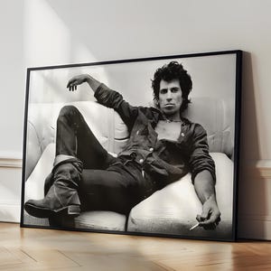 May include: Black and white photograph of a man sitting on a sofa, wearing a jacket and jeans. The photo is framed in a black frame.