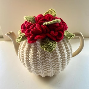 May include: A cream-coloured teapot with a knitted cosy and a large, red crocheted flower with green leaves on top. The teapot has a curved handle and spout.