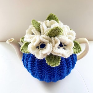 May include: A ceramic teapot with a blue knitted cosy, adorned with a cluster of white and green knitted flowers. The teapot features a white spout and handle. The cosy is a vibrant blue, and the flowers provide a decorative element.