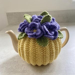 Könnte beinhalten: Eine cremefarbene Teekanne mit einer gelben Strickhülle. Die Hülle ist mit einem Cluster aus lila gestrickten Blumen und grünen Blättern bedeckt. Die Teekanne hat einen gebogenen Griff und einen Ausguss.