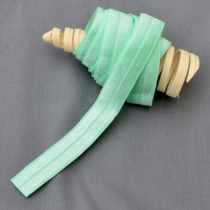 May include: A roll of light green elastic with a white stripe down the center. The elastic is rolled up on a wooden dowel.