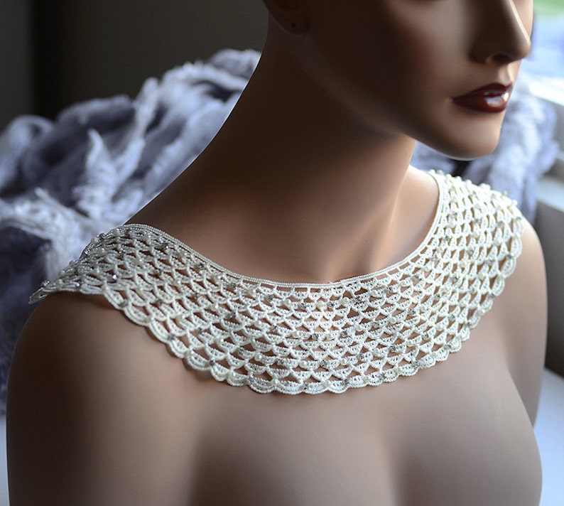 May include: A white crocheted collar with pearl-like beads and rhinestones. The collar is a delicate and intricate piece of jewelry.