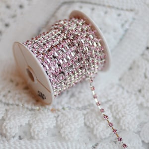 May include: A spool of silver chain trim with pink rhinestones. The chain is on a white background.