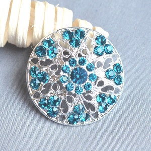 May include: A silver filigree brooch with a circular design featuring a large turquoise blue rhinestone center surrounded by smaller turquoise blue rhinestones.