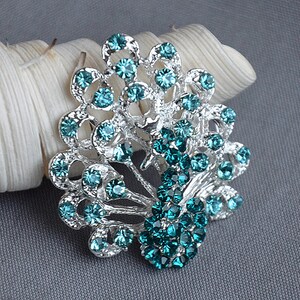 May include: A silver peacock brooch with teal blue rhinestones. The brooch is detailed with a peacock's tail feathers and a peacock's head.