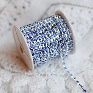 May include: A spool of silver chain with light blue rhinestones. The chain is coiled on a white spool.
