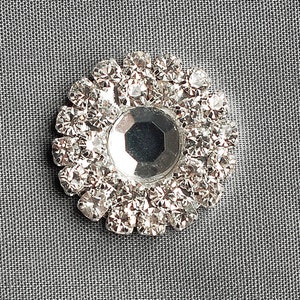 May include: A silver-toned, round, decorative embellishment with a central, faceted, clear crystal surrounded by smaller, clear crystals.
