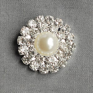 May include: A white pearl surrounded by a circle of clear rhinestones, forming a decorative embellishment.