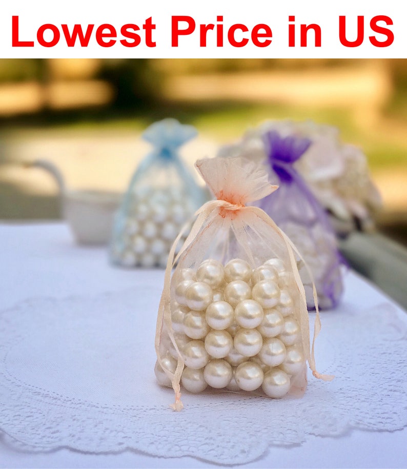 May include: A close-up of three small white organza bags filled with white pearl beads. The bags are on a white lace tablecloth. The text "Lowest Price in US" is at the top of the image.