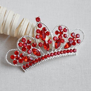 May include: A silver crown with red rhinestone accents. The crown is a decorative piece with a pointed top and a rounded base.
