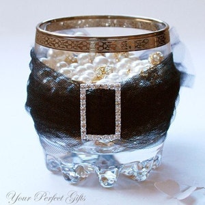 Pode incluir: Um vaso de vidro transparente com uma borda dourada e um laço de tule preto. O vaso está cheio de pérolas brancas e strass.