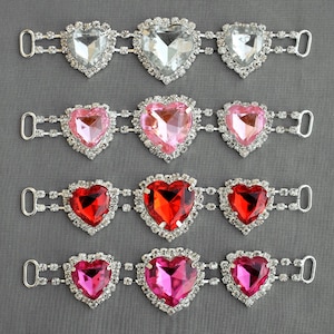 Puede incluir: Cinco filas de adornos de corazón de pedrería con cierres de metal plateado. Las pedrerías son transparentes, rosas, rojas y fucsias.