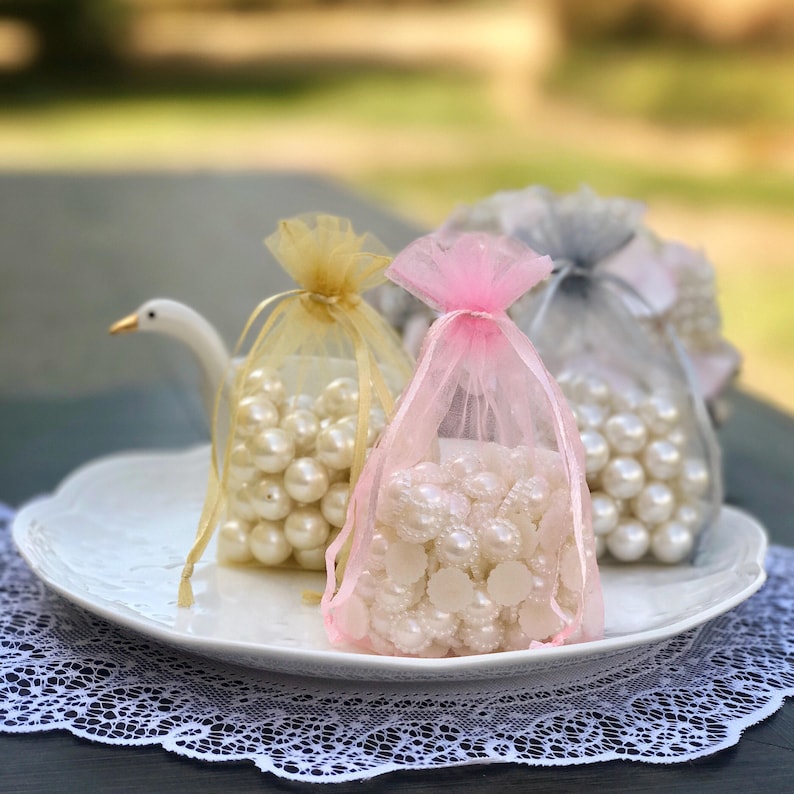 May include: Three small bags filled with white pearl beads. The bags are made of sheer fabric in pink, gold, and gray. The bags are sitting on a white plate.