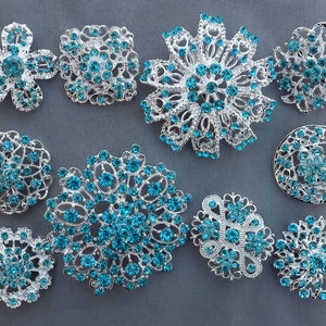May include: Assortment of 10 silver brooches with turquoise blue rhinestones. The brooches are in various shapes, including flowers, circles, and geometric designs.