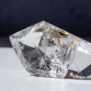 May include: A clear quartz crystal with sharp, faceted edges. The crystal has internal inclusions of gold and brown, and sits on a white surface against a dark background. The crystal is a natural, raw gemstone.
